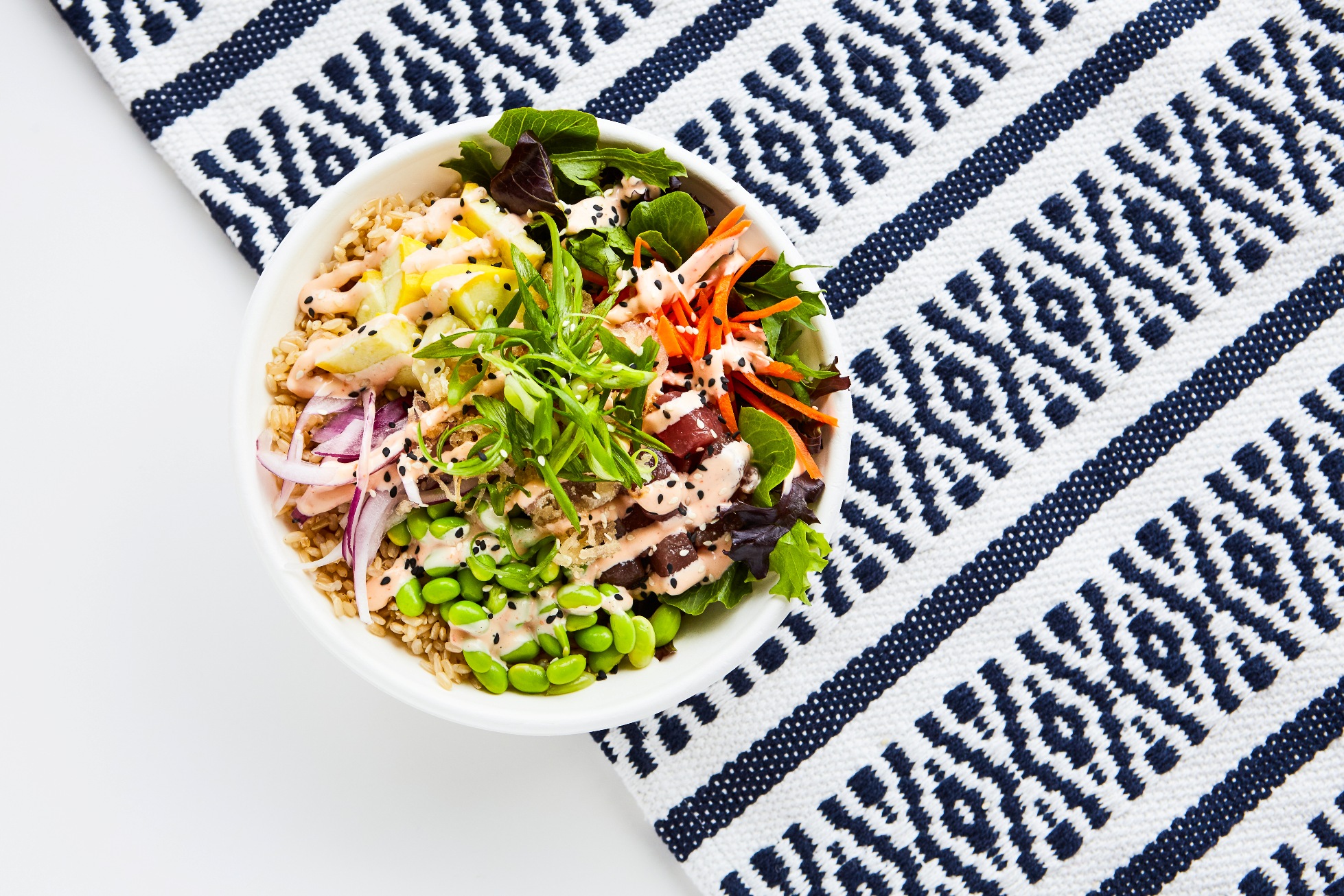 Poke bowls with fresh ingredients including tuna &amp; chicken with fresh vegetables sauced up in BPI certified compostable bowls on a white table with a navy blue printed graphic runner.